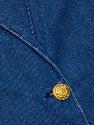 Close-up of the Baldwin Blazer by L'AGENCE, featuring blue indigo wash denim, a gold textured button, and visible white stitching along the fabric edge.