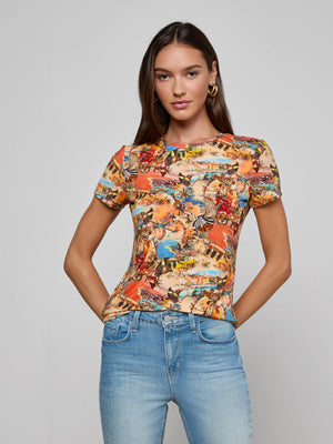 A woman with long brown hair wears the L'AGENCE Ressi Fitted Tee, featuring a colorful collage print in stretch jersey knit, paired with light blue jeans. She stands against a plain light gray background, looking at the camera with her hands in her pockets.