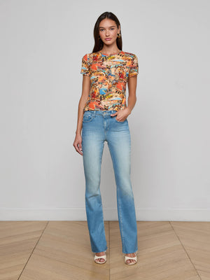 A woman with straight brown hair and a relaxed expression stands indoors against a white wall, wearing the L’AGENCE Ressi Fitted Tee in travel collage print, light blue bootcut jeans, and white open-toe heels with one hand in her pocket.