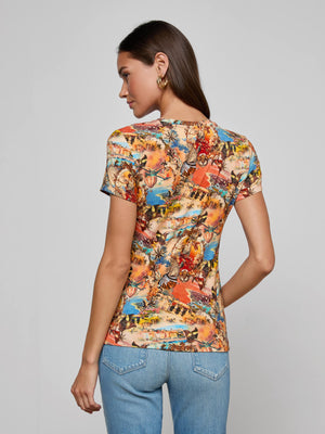 A woman with long brown hair, wearing gold hoop earrings, a L’AGENCE Ressi Fitted Tee with a travel collage print, and light blue jeans stands facing away against a plain light gray background.