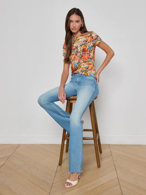 A woman with long brown hair sits on a wooden stool against a plain white wall, wearing the L'AGENCE Ressi Fitted Tee, light blue jeans, and beige heeled sandals on a light wood floor.