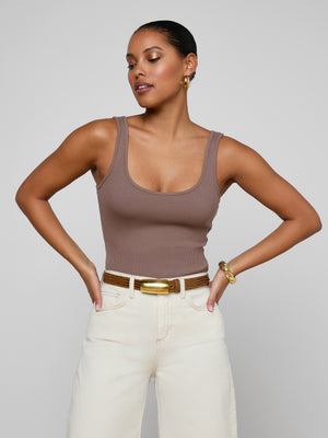 Wearing the Nica Scoopneck Bodysuit, a woman pairs it with cream pants, a brown belt, gold hoop earrings, and a bracelet, confidently posing with hands on hips against a plain light background.
