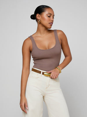 A woman with dark hair in a low bun wears the mauve Nica Scoopneck Bodysuit and high-waisted cream pants, accessorized with gold hoop earrings, a bracelet, and a brown belt, posing against a plain light gray background.