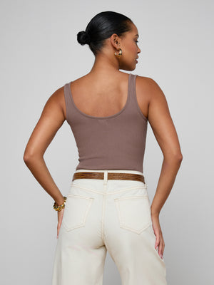 A woman stands facing away, wearing the sculpted fit Nica Scoopneck Bodysuit in brown with high-waisted cream pants and a brown belt. Her hair is styled in a bun, accessorized with gold hoop earrings and a bracelet.