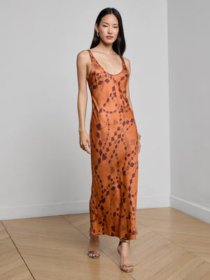 A woman with long, straight hair wears the L'AGENCE Akiya Satin Maxi Dress—a burnt orange, bias-cut dress with a geometric pattern. She pairs it with heeled sandals and poses in a minimal room, one hand relaxed by her side.
