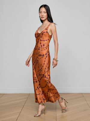A woman in the L'AGENCE Akiya Satin Maxi Dress stands on a wooden floor against a light gray wall. The bias-cut burnt orange dress features a geometric pattern. She pairs it with gold heels, bracelets, earrings, and wears her long hair straight.