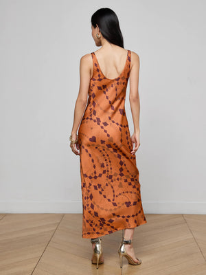 A woman with long dark hair wears the L'AGENCE Akiya Satin Maxi Dress, standing with her back to the camera in a minimalist room with light wood flooring.