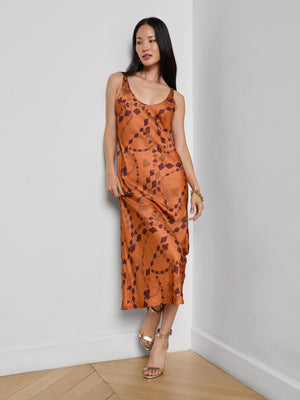 A woman in the Akiya Satin Maxi Dress by L'AGENCE stands on hardwood floors, wearing gold sandals and a bracelet. She has long straight hair and a relaxed expression. The dress is burnt orange with a bias-cut tank silhouette.