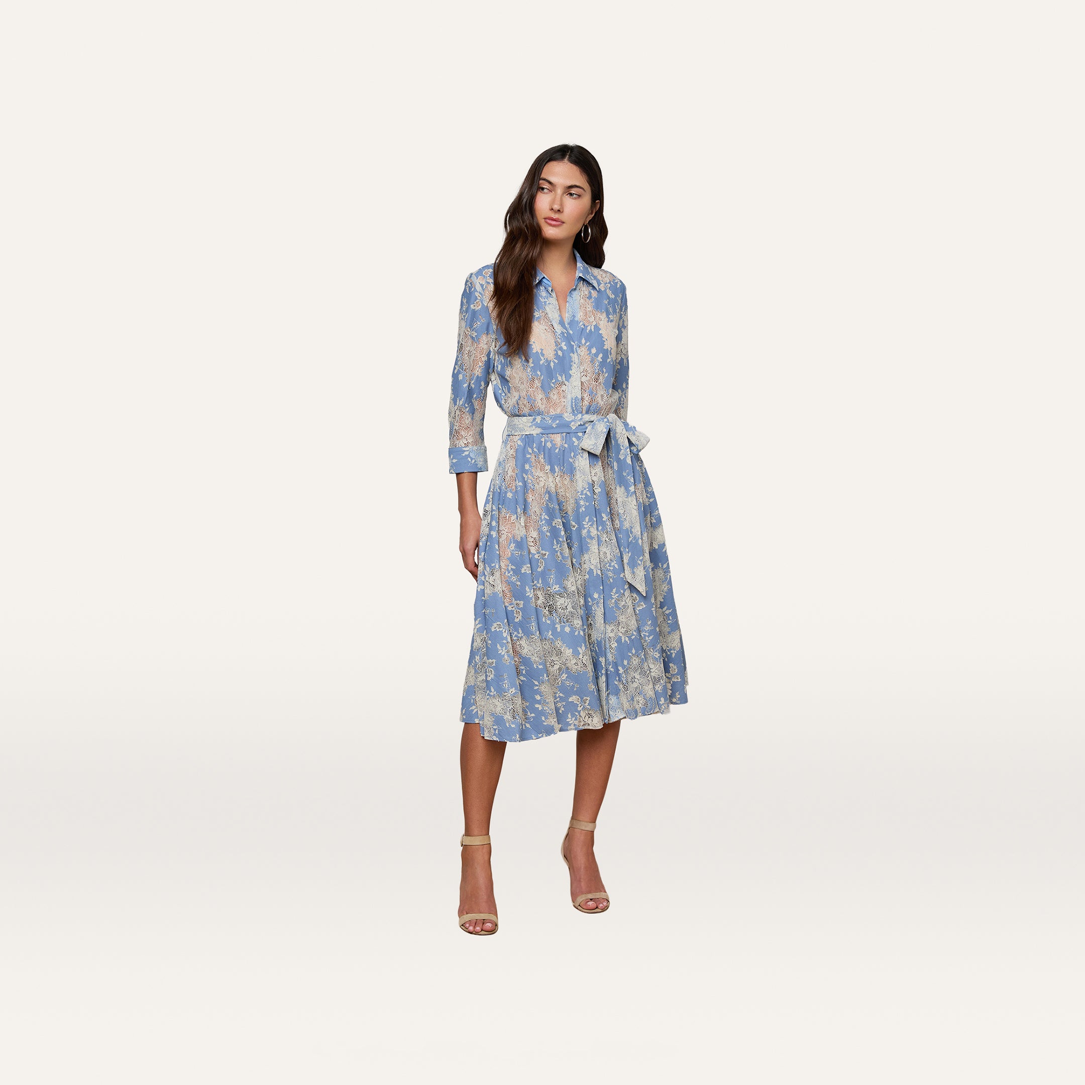 Woman wearing the Ralphie Lace Midi Dress in light blue, featuring long sleeves, a knee-length silhouette, floral lace detailing, and a tied waist; styled with beige heels against a plain white background.