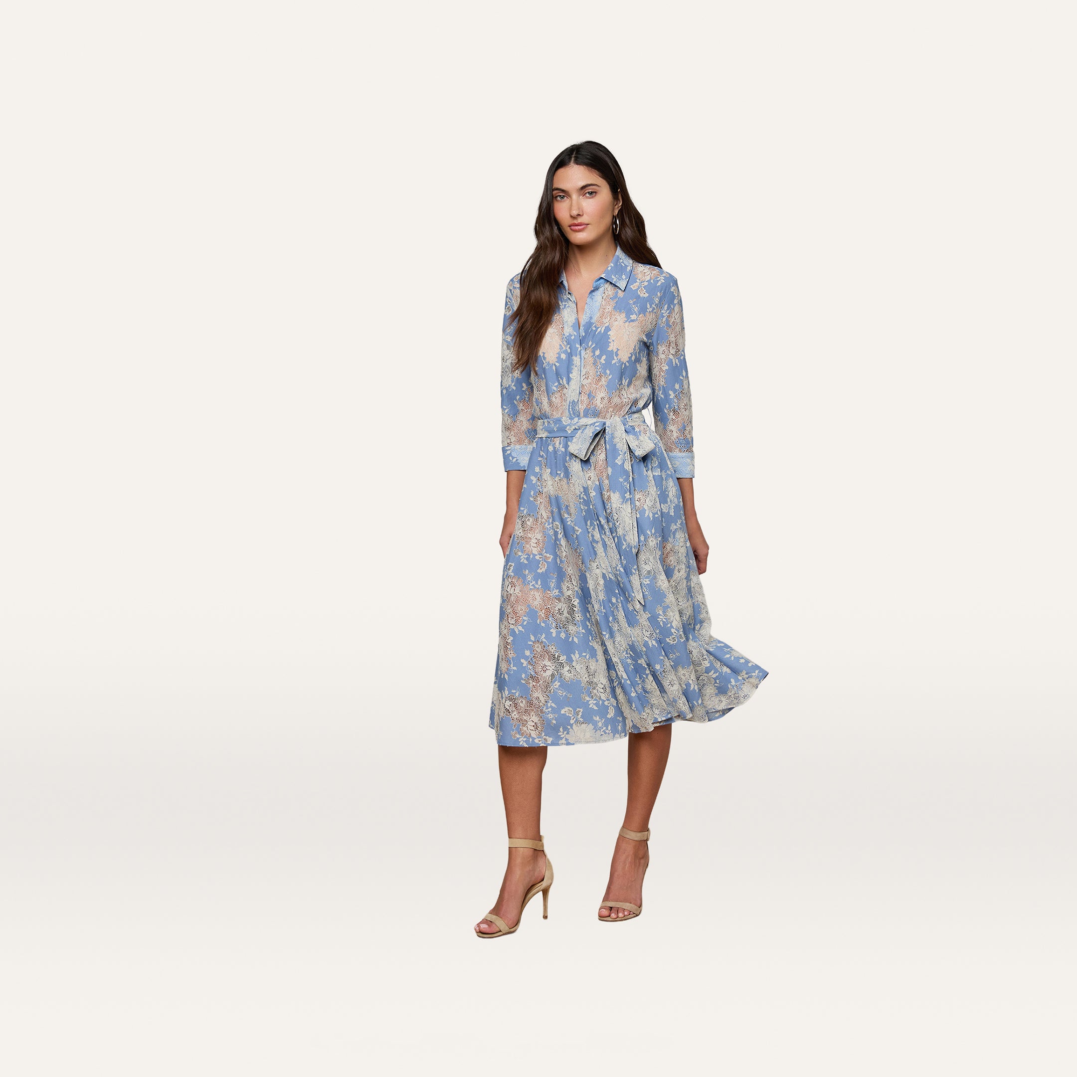 A woman wearing a light blue and beige floral Ralphie Lace Midi Dress with long sleeves and a tie belt, paired with beige high-heeled sandals, stands against a plain white background.