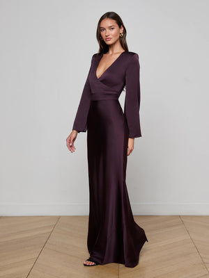 A woman stands on a light wooden floor against a plain white wall, wearing the Manon Satin Gown—a deep purple, floor-length gown with long sleeves and a plunging neckline.