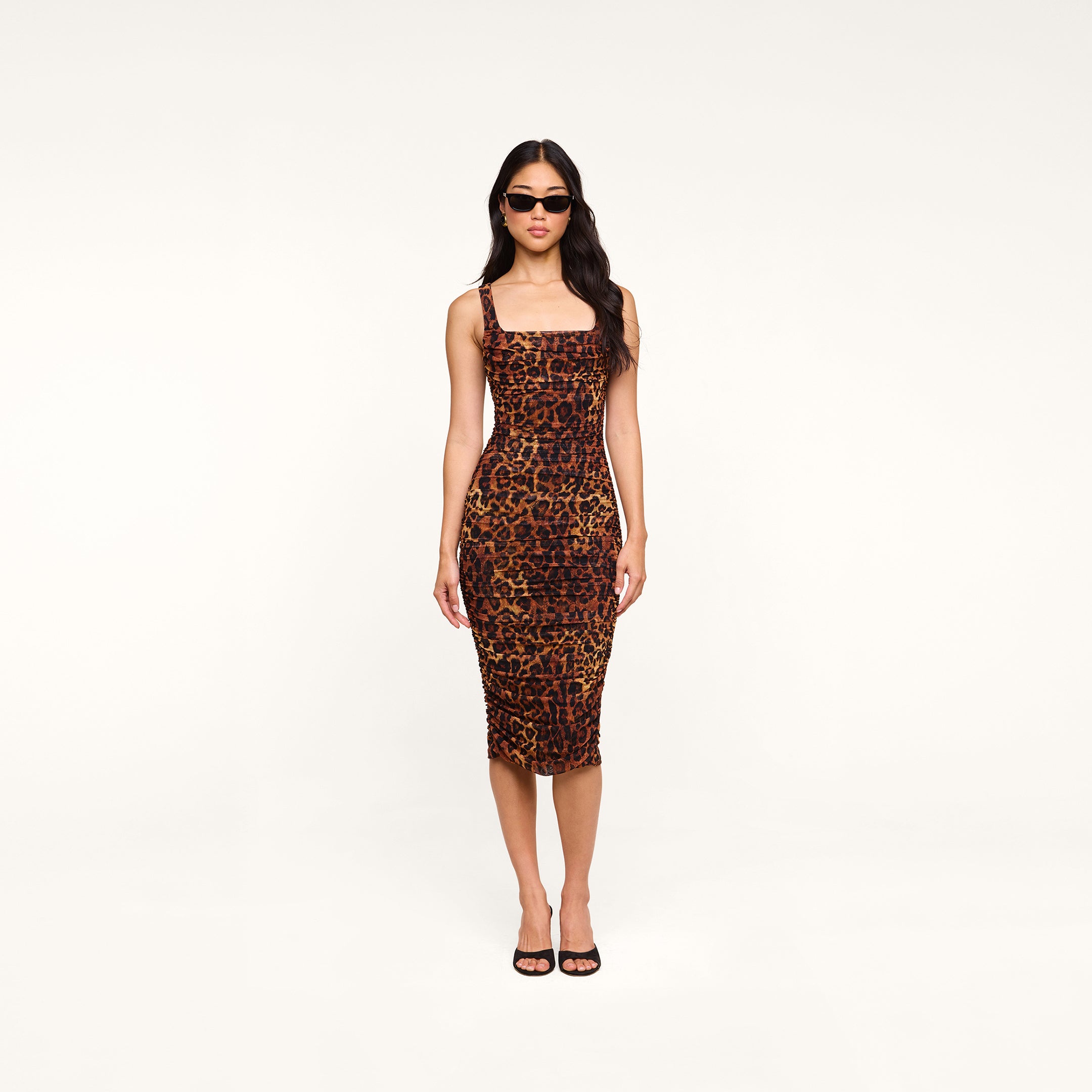 A woman stands facing forward with her arms relaxed at her sides, wearing the Anya Shirred Dress—a form-fitting leopard print midi dress—paired with black sunglasses and black open-toe heels. The background is plain white.
