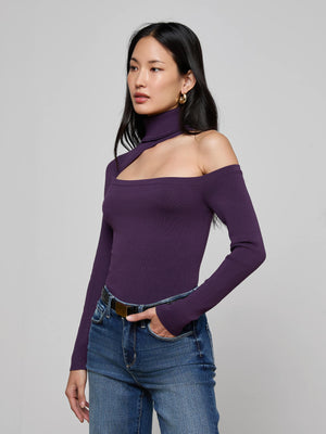 A woman with long dark hair and gold hoop earrings wears the L'AGENCE Azia Cutout Knit Top—an asymmetrical purple off-shoulder turtleneck—paired with blue jeans, standing against a plain light gray background with a neutral expression.