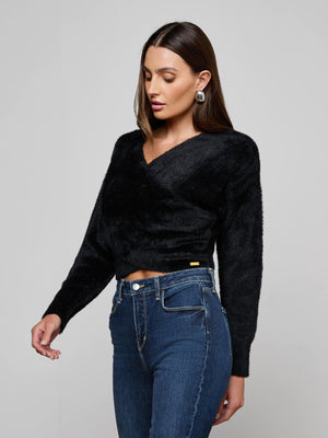 A woman with straight brown hair and large round earrings wears the L'AGENCE Antonella Crossover Sweater with high-waisted blue jeans, standing against a gray background and looking down slightly with one arm by her side.