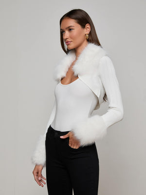 A woman with long brown hair wears an ivory fitted top and black high-waisted pants, styled with the L'AGENCE Haru Faux Fur Shrug—a cropped jacket featuring removable faux fur collar and cuffs—channeling old Hollywood glamour against a plain light background.