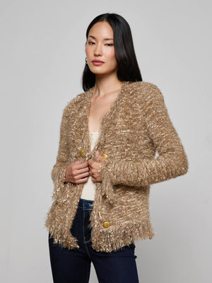 A woman with long dark hair wears the L'AGENCE Azure Fringe Cardigan Blazer, a textured gold-beige knit with fringe trim and large gold buttons, over a light top and dark jeans against a plain gray background.