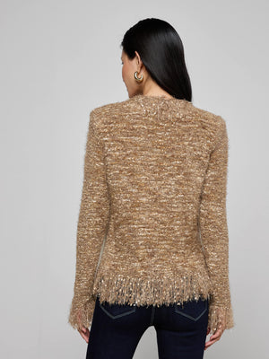 A woman with long dark hair and hoop earrings models the L'AGENCE Azure Fringe Cardigan Blazer, a gold-brown knit with fringe trim at the hem and cuffs, paired with dark jeans, as she stands with her back to the camera.