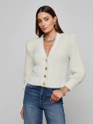 A woman with straight brown hair wears the L'AGENCE Talulla Cardigan, a fuzzy white button-up with gold buttons, paired with blue jeans and gold jewelry, standing against a plain light gray background.
