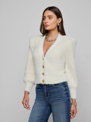 A woman wearing the white Talulla Cardigan by L'AGENCE with gold buttons, blue jeans, gold earrings, a chunky gold necklace, and a gold watch stands against a plain gray background.
