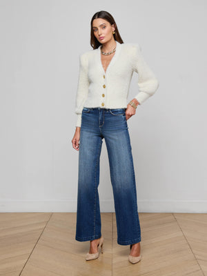 A woman poses indoors against a light wall, wearing the L’AGENCE Talulla Cardigan in white, paired with blue wide-leg jeans, beige heels, and jewelry. She stands on a wooden floor with one hand on her hip.