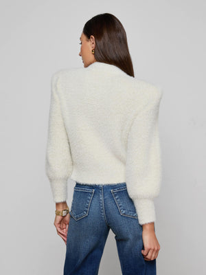 A woman with long brown hair, wearing the L'AGENCE Talulla Cardigan—a fuzzy white knit with puffed sleeves—paired with blue jeans, stands facing away from the camera against a plain light gray background.