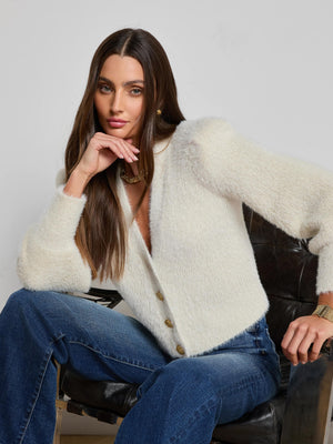 A woman with long brown hair wears the L'AGENCE Talulla Cardigan in white and blue jeans as she sits on a black chair, confidently resting her chin on her hand and looking at the camera.