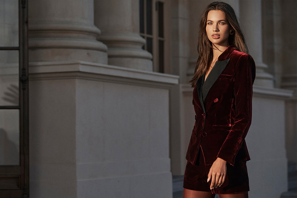 A woman stands outside a building wearing a burgundy velvet blazer and matching shorts, with sunlight illuminating her face and the columns behind her. The soft velvet material adds richness to her look as she confidently poses in the warm glow.