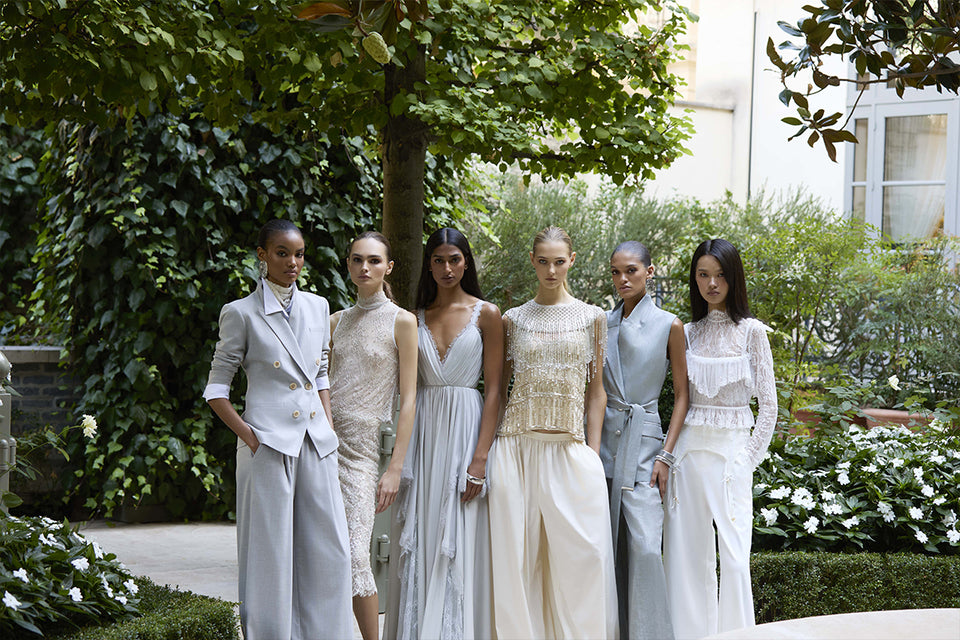 Six women in elegant, light-colored gowns and suits stand together outdoors in a lush garden with vibrant flowers, posing stylishly under a tree for a chic fashion photo.
